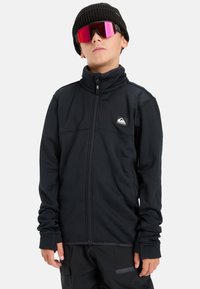 Czarna kurtka na zamek z wysokim kołnierzem, teksturowana tkanina i naszywka z logo. Noszona z czapką beanie z dzianiny w kolorze czarnym oraz różowymi, lustrzanymi okularami przeciwsłonecznymi.