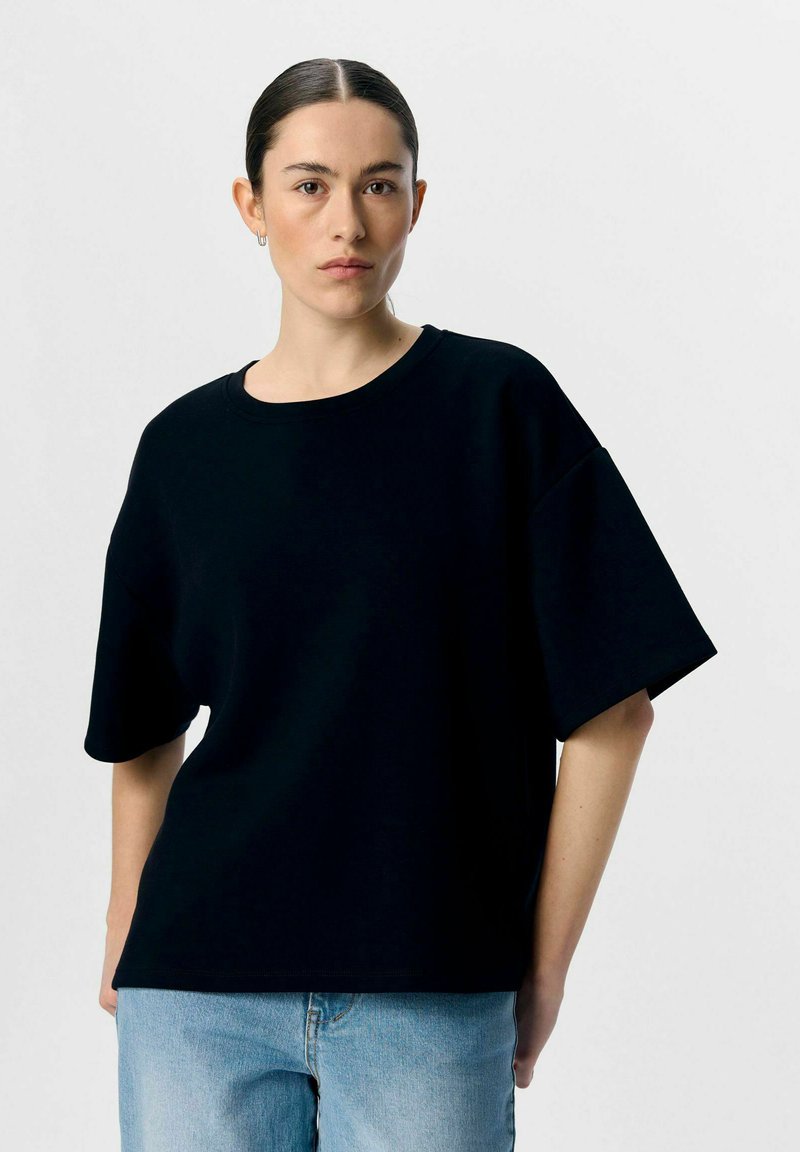 Zwarte oversized t-shirt met korte mouwen, ronde halslijn en een zachte textuur. Gecombineerd met lichtblauwe spijkerbroeken. Neutrale achtergrond.