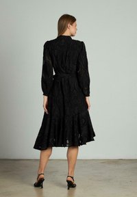 Schwarzes Midikleid mit ausgestelltem Rock, das mit aufwendiger Blumenstickerei und einem taillierten Bund versehen ist. Lange Ärmel und ein hoher Ausschnitt.