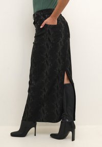 Jupe maxi en imprimé serpent noir avec fente sur le côté et poches ; associée à de hautes bottes à talons aiguilles noires ; tissu lisse et texturé.
