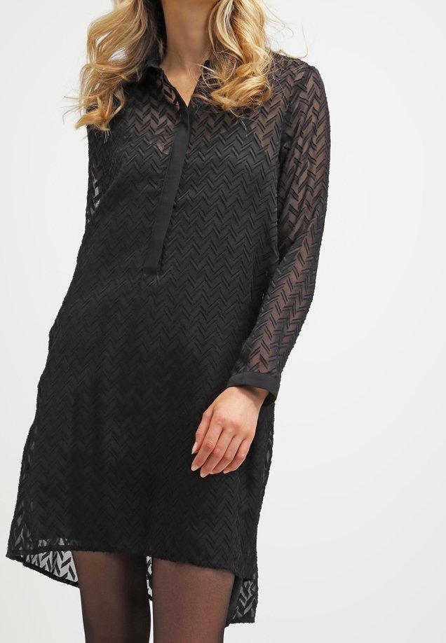 Femme portant une robe noire transparente texturée à manches longues avec des motifs en chevrons et un col, debout avec une main près de la cuisse.