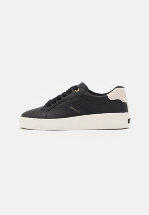 Zapatilla de cuero negro con suela de goma blanca, con un detalle beige en el talón, punta redonda y cordones negros. Detalle de logo dorado en el lateral.