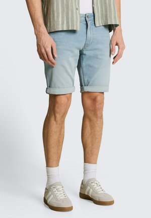 Man draagt lichtblauwe opgerolde denimshorts, witte sokken, beige sneakers en een gestreept overhemd met zichtbare knopen.