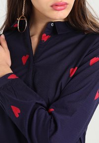 Marineblaues Hemd mit Kragen, verziert mit roten Herzmustern. Langärmelig mit geknöpften Manschetten, aus leichtem Stoff mit glatter Textur.