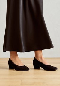 Svarta mockaskor med låg klack och scallopmönster, kombinerade med en flödande mörk klänning, på en ljus träplankegolv.