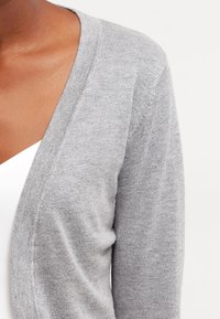 Ljusgrå cardigan med V-ringning, mjuk textur och något öppen fram, som visar den släta tyget och konturen av en vit topp under.