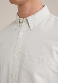 Chemise boutonnée gris clair avec de subtiles rayures verticales, un col classique et une poche poitrine unique. Tissu doux avec une finition texturée.