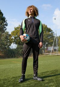 Chándal atlético negro con acentos en verde neón. De pie sobre el césped, sosteniendo un balón de fútbol, vistiendo botas de fútbol negras. Fondo de cielo despejado.