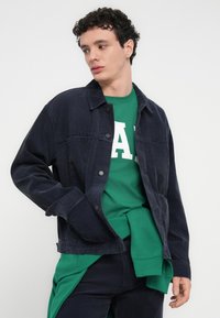 Μπλε μπουφάν από κορδόνι με άνετη εφαρμογή, με μαύρα κουμπιά και πλαϊνές τσέπες, φορεμένο πάνω από πράσινη φούτερ μπλούζα με μεγάλο λευκό γράμμα.