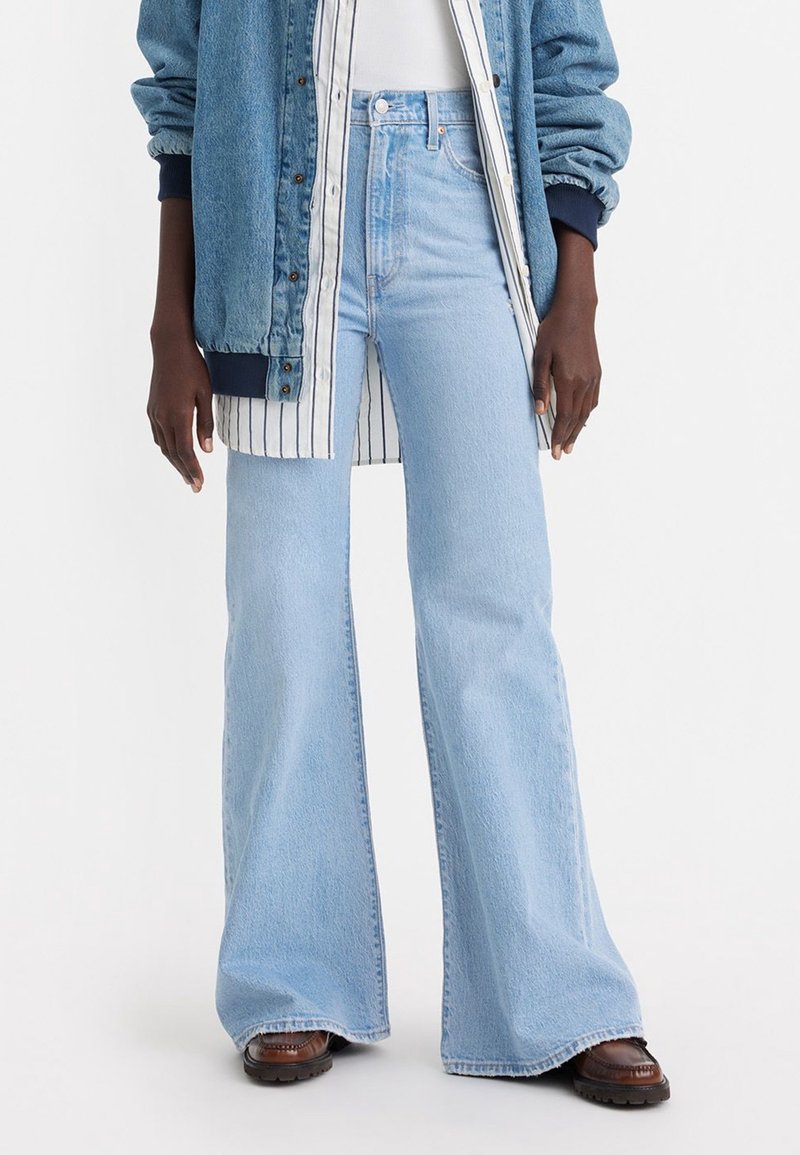 Personne portant un jean bleu clair taille haute évasé, des chaussures marron, une chemise blanche, une chemise à rayures boutonnée et une veste en jean bleue avec des poignets élastiques.