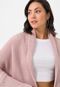 Weiche, rosa gestrickte Strickjacke mit gerippter Textur, offener Front und weiten Ärmeln, darüber getragen ein weißes, geripptes Crop-Top.