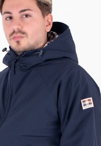 Giacca a vento navy con zip frontale, cappuccio regolabile con coulisse e fodera interna a quadri. Presenta una toppa con logo sulla manica sinistra.