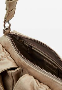 Sac à main beige avec une surface extérieure douce et texturée. L'intérieur comprend une poche zippée et une doublure en satin. Quincaillerie métallique et bandoulière réglable visibles.