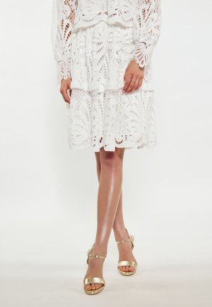Rochie albă din dantelă, cu decupaje florale, design stratificat și mâneci lungi. Pantofi cu toc aurii, cu curele, completează ținuta. Tonul pielii este neutru.