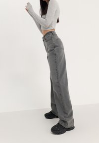 Vaqueros grises de pierna ancha con cintura alta, hechos de mezclilla. Presentan un acabado limpio y bolsillos laterales. Combinados con zapatillas negras de punta redondeada.