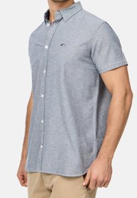 Camisa de botones de manga corta en azul claro, con una textura sutil, un cuello, botones en la parte delantera y un logo bordado en el pecho.