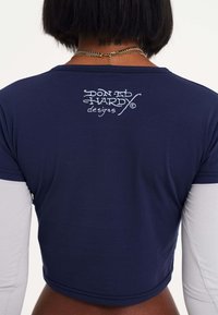 Cropped top navy con maniche lunghe grigie, con stampa del logo bianco sul retro. Realizzato in tessuto morbido e elastico con un design aderente.