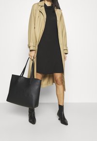Beiger Trenchcoat über einem schwarzen Kleid, kombiniert mit einer großen schwarzen Tragetasche und schwarzen Ankle Boots. Glatte Textur; einfaches Design.