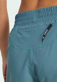 Türkise Laufshorts mit elastischem Bund, ausgestattet mit einer Außentasche mit einem schwarzen Reißverschluss. Glatte Textur und Flachnähte.