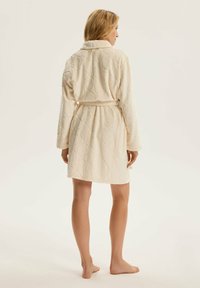 Kremowy szlafrok wykonany z miękkiej, pikowanej tkaniny. Posiada kołnierz, pasek w talii oraz długie rękawy.