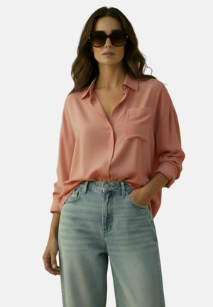 Vrouw met lang golvend haar die oversized donkere zonnebril, loszittend roze overhemd met knopen en lichtblauwe hooggetailleerde spijkerbroek draagt, met één hand in de zak.