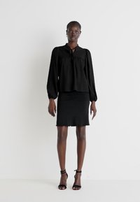 Svart bluse med lange, puffete ermer og knytehals, paret med et tettsittende svart skjørt. Begge antrekkene har et mykt, teksturert stoff.