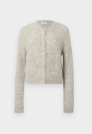 Femme aux cheveux foncés portant un cardigan beige duveteux boutonné, un jean large taille haute foncé avec ceinture, et des chaussures plates marron, debout les mains dans les poches.