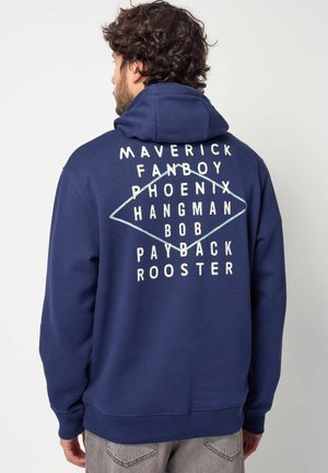Sudadera azul marino con capucha ajustable; presenta texto blanco y un diseño geométrico en la parte posterior. El material parece suave y texturizado.