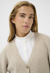 Beige strikket cardigan med V-hals, med en træknap, båret over en hvid skjorte med flæser. Glat tekstur og afslappet design.