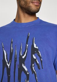 Camiseta de algodón azul con un gran estampado gráfico negro y acentos blancos. Cuello redondo y mangas cortas, tela texturizada.