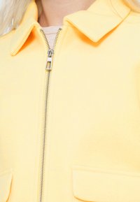 Μπουφάν μίγματος μαλλιού σε κίτρινο χρώμα με φερμουάρ μπροστά, κλασικό γιακά και δύο μπροστινές τσέπες. Μαλακή υφή και εφαρμοστή γραμμή.