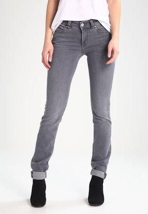 Personne portant un jean gris slim-fit retroussé aux chevilles, une chemise blanche et des bottines noires, debout devant un fond uni.