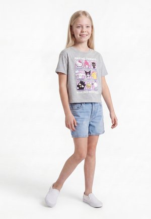 Fille souriante aux longs cheveux blonds, portant un t-shirt gris avec un imprimé cartoon, un short en jean bleu clair et des chaussures blanches sans lacets.