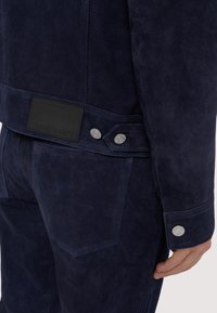 Navy modrý kordurový kabát s hladkým povrchem, stříbrnými knoflíky a černou koženou nášivkou na zadní straně.