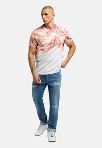 Grafisk t-skjorte med rosa og kremfargede svimerker, hvit nedre del og rød tekst. Brukes med slitte blå jeans og hvite sneakers.