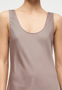 Άτομο που φοράει αμάνικο τοπ ταουπ με στρογγυλή λαιμόκοψη, εμφανίζοντας το πάνω μέρος του κορμού και μερικά τατουάζ στα εσωτερικά μπράτσα έναντι απλού φόντου.