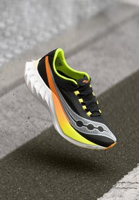 Baskets noires et grises avec une tige texturée, accents verts, bande orange et design de semelle distinctif. Comprend six œillets jaunes et des lacets.