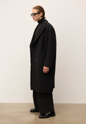 Persoon in een zwart oversized dubbelrijgjas, zwarte coltrui, wijdvallende broek, glanzende zwarte loafers en donkere zonnebril, staand in zijaanzicht.