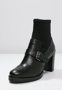 Botín negro de tacón alto, hecho de cuero suave y tejido elástico. Presenta una correa con hebilla, un intrincado detalle perforado y un tacón grueso.