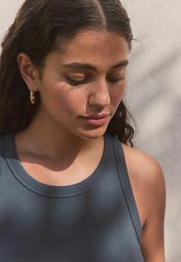 Donkergrijze geribbelde tanktop met een ronde halslijn en een gestructureerde afwerking. Model draagt kleine gouden ringoorbellen en heeft golvend donkerhaar.