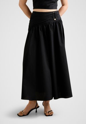 Femme portant une longue jupe noire avec une ceinture à boucle dorée et des sandales à talons ouvertes noires avec des accents dorés.