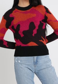 Donna che indossa un maglione aderente con motivi astratti neri, arancioni e rosa abbinato a jeans di denim a vita alta di un blu chiaro.