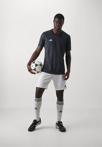 Vīrietis futbolists melnā krekla un baltajās biksēs tur futbolu, stāvot pret vienkāršu baltu fona.