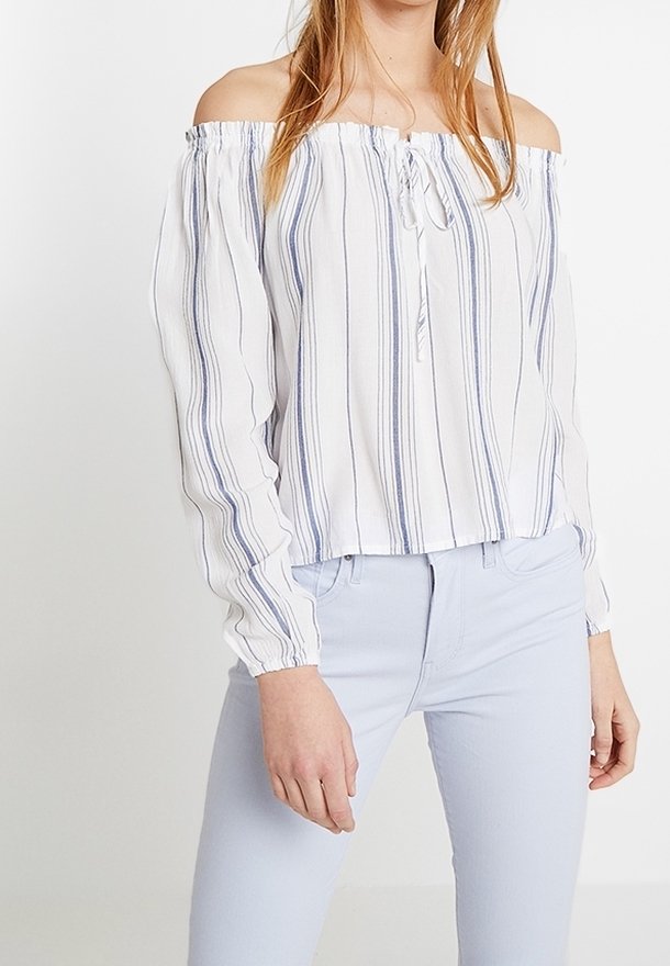 Femme portant un chemisier blanc à épaules dénudées avec des rayures verticales bleues et un pantalon ajusté bleu clair sur un fond uni.