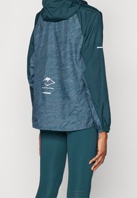 Teal atletisk jakke med en struktureret, mønstret ryg, justerbar hætte og reflekterende detaljer. Sammen med matchende leggings med udskæringer.