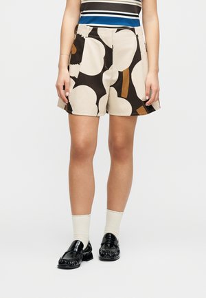 Nő bézs rövidnadrágban, melyen nagy fekete és barna absztrakt minták találhatók, fehér zoknival és fekete mokaszinokkal párosítva.