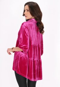 Top in velluto rosa con design a strati, dotato di maniche a tre quarti e orlo asimmetrico, indossato con leggings neri e braccialetti.
