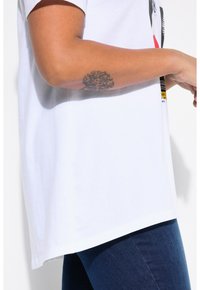 Camiseta de algodón blanca con un corte relajado, con un diseño impreso en negro, rojo y amarillo. Vaqueros de mezclilla azules visibles debajo de la camiseta.