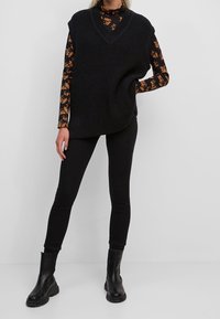 Gilet oversize a coste nere con scollatura a V, indossato sopra un dolcevita a maniche lunghe con stampa floreale. Abbinato a jeans skinny neri e stivali alla caviglia.