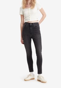 Svarta högmidjade jeans med smal passform och fickor framtill, kombinerat med en vit ruggad crop top och beige sneakers.